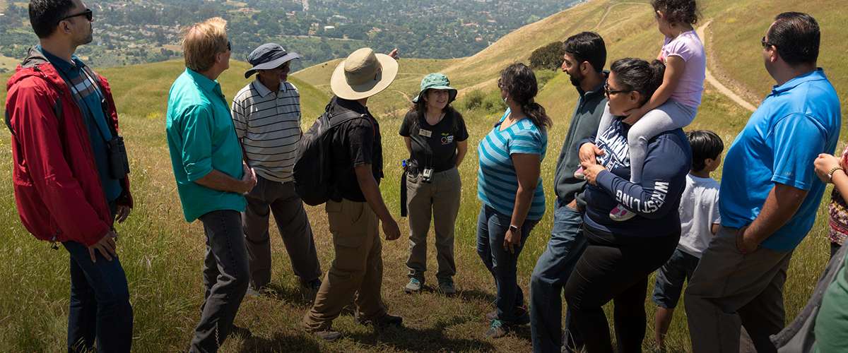 Group of 12 adults and children taking in the view at the top of Sierra Vista, surrounding a female volunteer in an Open Space Authority t-shirt